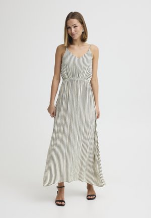 Vrouw draagt een mouwloze gestreepte maxi-jurk met dunne bandjes en zwarte open schoenen, staande tegen een effen lichte achtergrond.