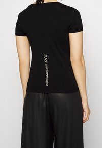 Camiseta negra de manga corta con un diseño ajustado. Presenta el texto blanco "EA7 Emporio Armani" a lo largo de la parte posterior para la marca. Textura de tela suave.