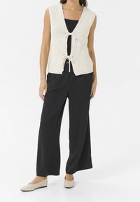 Kvinde iført ærmeløs hvid strikket vest bundet foran over en sort top, brede sorte bukser og beige flade sko, stående foran en hvid baggrund.