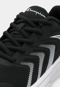 Fekete sportcipő textúrált anyaggal, fehér részletekkel és jellegzetes csíkozással az oldalon, fekete fűzőkkel és párnázott gallérral.