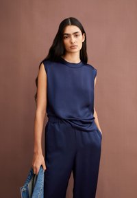 Navy blue, sleeveless top with a smooth, satin finish, paired with loose pants. The outfit features a subtle draped detail and pockets.