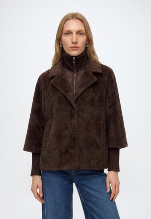 Femme aux longs cheveux blonds porte un manteau en fausse fourrure marron foncé par-dessus un pull à col roulé marron zippé et un jean bleu, debout devant un fond uni.