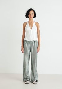 Bílý halter top se zipem, doplněný zelenými pruhovanými kalhotami s širokými nohavicemi. Modelka má na sobě černobílé boty. Jednoduché, minimalistické pozadí.