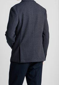 Man som bär en mörk strukturerad blazer och marinblå byxor, sedd bakifrån mot en ljus, enkel bakgrund.
