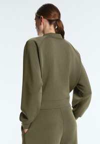 Femeie cu păr castaniu prins în coadă de cal, purtând un top cu mâneci lungi cu dungi și pantaloni asortați de culoare verde măsliniu, cu mâinile în buzunare, orientată lateral.