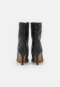 Botines negros de tacón alto con patrón perforado, cierre de cremallera, textura suave y silueta elegante. Logo del diseñador visible en el interior.