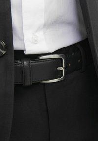 Ceinture en cuir noir avec une boucle en métal argenté, portée sur un pantalon noir et une chemise blanche. Détails de couture visibles et bouton.