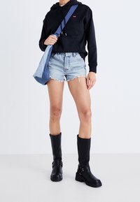 Felpa nera con cappuccio e logo rosso, pantaloni corti in denim blu chiaro con orli sfrangiati e stivali neri in pelle alti con fibbie e suola spessa.