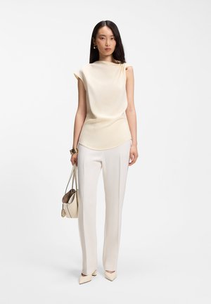 Femme debout portant un haut crème sans manches, un pantalon blanc sur mesure, des talons beige, des bracelets en or, des boucles d'oreilles et tenant un sac à main beige.