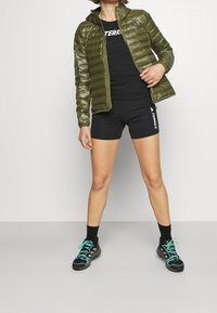 Osoba w oliwkowej, pikowanej kurtce, czarnych szortach i bluzie marki Terrex oraz czarnych trampkach z turkusowymi sznurowadłami na jednolitym tle.