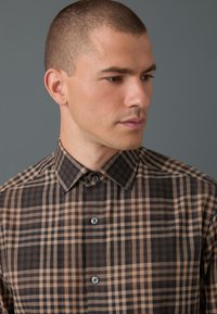 Camicia a quadri marrone e beige con colletto abbottonato, caratterizzata da bottoni normali e una texture liscia. Il motivo è composto da quadretti di varie dimensioni.