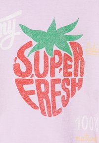 Heled lilla T-särk, millel on suur graafika punasest maasikast roheliste lehtedega. Tekst ütleb "SUPER FRESH" ja "100% looduslik."