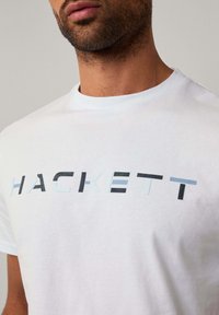 Camiseta de algodón blanca con un audaz logo "HACKETT" en diferentes tonos de azul y negro. Mangas cortas y cuello redondo.