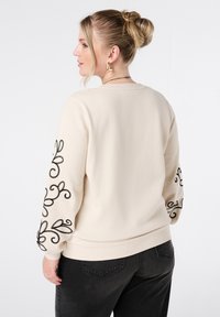 Sweat-shirt beige avec broderie noire sur les manches, col rond, poignets et ourlet côtelés. Porté avec un jean gris foncé. Texture lisse.