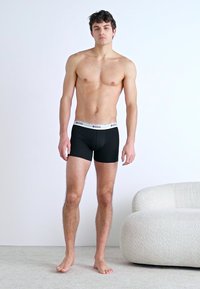 Schwarze Boxershorts mit einem weißen Bund, auf dem "BOSS" steht. Ein männliches Model mit kurzen Haaren steht barfuß vor einem schlichten Hintergrund.