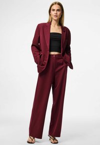 Burgundy kostym med avslappnade byxor och en matchande blazer, stylad över en figurnära svart crop top. Slätt tyg med strukturerad design.