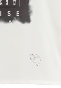 Camiseta blanca con una impresión gráfica negra que presenta texto estilizado y un pequeño acento de corazón de strass en la esquina inferior derecha.