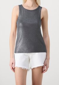 Kvinna i en metallisk grå ärmlös topp och vita slitna denimshorts, stående mot en enkel ljus bakgrund.