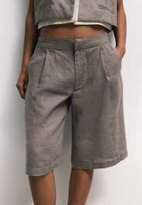 Persona que lleva unos pantalones cortos de lino gris sueltos, de largo hasta la rodilla, con pliegues y bolsillos laterales, combinados con un chaleco corto a juego.