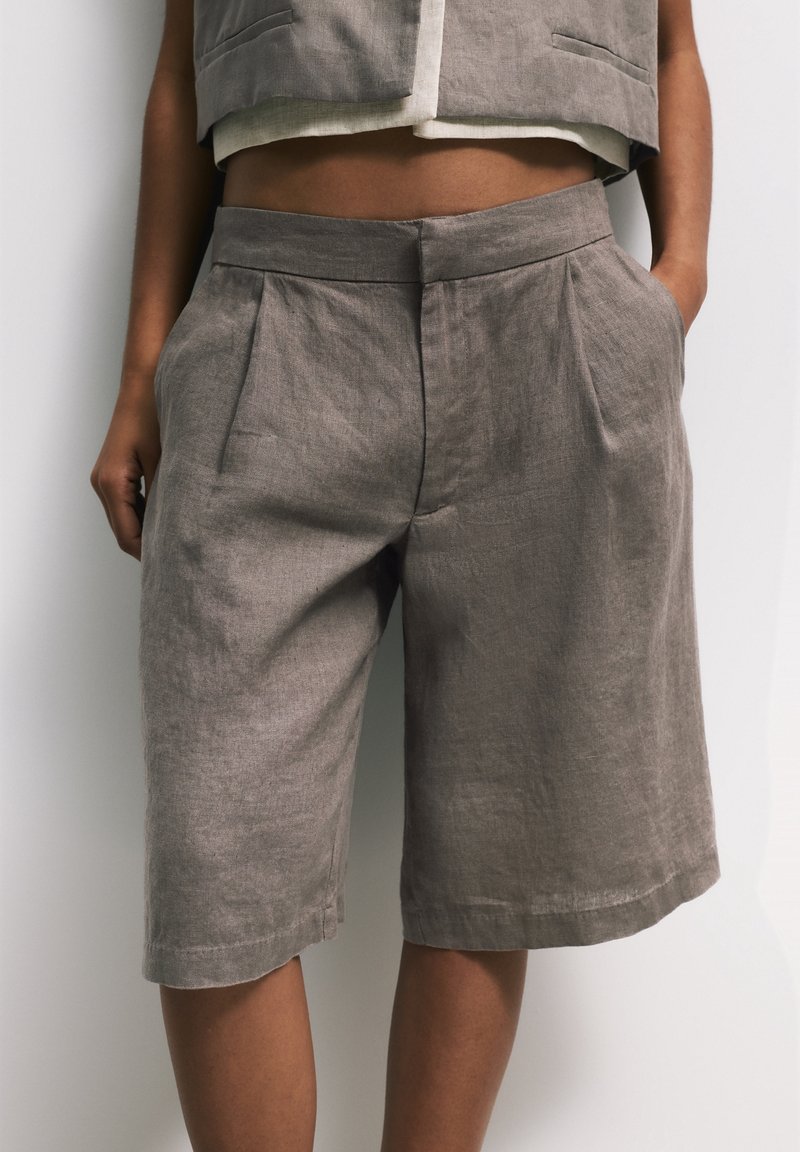 Persona que lleva unos pantalones cortos de lino gris sueltos, de largo hasta la rodilla, con pliegues y bolsillos laterales, combinados con un chaleco corto a juego.