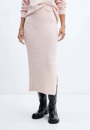 Femme portant une jupe midi en tricot côtelé rose pâle et un pull assorti avec des bottes en cuir noires mi-mollet sur un fond uni.