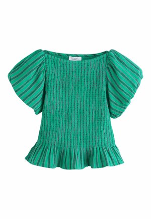 Frau trägt ein türkis gestreiftes Oberteil mit Puffärmeln und passende weit geschnittene Hose, steht draußen in der Nähe einer weißen Steinmauer mit Bäumen im Hintergrund.