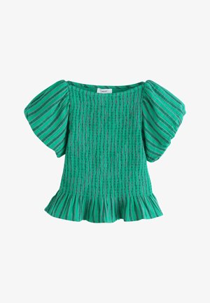 Kvinde iført tealstribet top med pufærmer og matchende vidde bukser, står udendørs nær en hvid stenmur med træer i baggrunden.