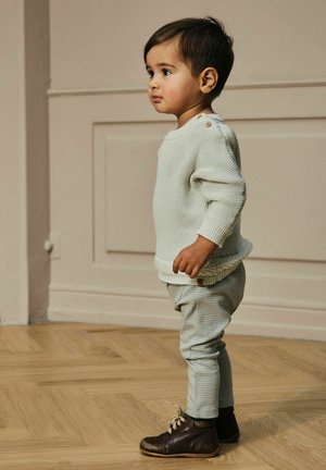 Peuterkind dat binnen op een houten vloer staat, gekleed in een crèmekleurige gebreide trui, gestreepte broek en bruine veterschoenen, kijkend naar de zijkant.