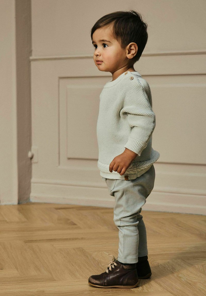 Peuterkind dat binnen op een houten vloer staat, gekleed in een crèmekleurige gebreide trui, gestreepte broek en bruine veterschoenen, kijkend naar de zijkant.