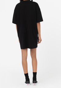 Osoba w czarnej, oversize'owej sukience t-shirtowej, czarnych skarpetach do połowy łydki i czarnych niskich sneakersach, stoi tyłem do kamery.