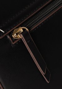 Angolo di una borsa in pelle nera con chiusura a zip, hardware dorato e un zip pull in pelle marrone a contrasto con due estremità piatte.