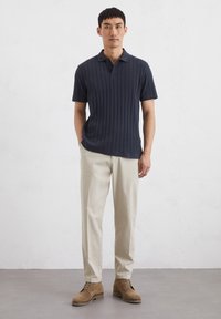 Marineblauwe geribbelde polo met korte mouwen, twee knopen bij de kraag; beige broek van lichte stof; bruine suède schoenen met veters.