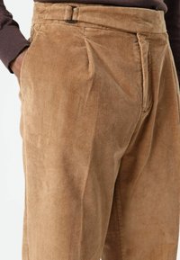 Tan corduroy trousers with vertical ribbing, featuring pleats and a waistband with a buckle. Smooth texture and tailored fit.