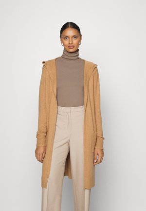 Long gilet couleur camel en matière douce, avec une capuche, une ouverture sur le devant et des poignets côtelés, porté sur un col roulé beige et un pantalon clair.