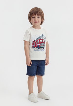 Νεαρός αγόρι με λευκά αθλητικά παπούτσια, τζιν σορτς και λευκό μπλουζάκι με κόκκινο πυροσβεστικό όχημα και την επιγραφή "Μελλοντικός Ήρωας", στέκεται και χαμογελάει.