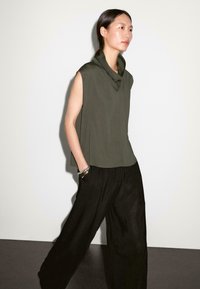 Canotta verde oliva senza maniche con scollo a drappeggio abbinata a pantaloni neri larghi. La modella si trova di fronte a uno sfondo chiaro e uniforme.