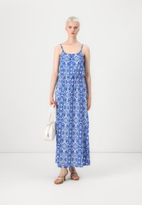 Blå och vit mönstrad maxikjol med tunna axelremmar, rynkad midja och flödande design. Matchad med en vit handväska och bruna sandaler.