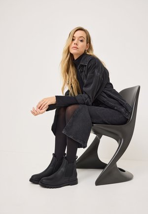 Chaqueta de denim negra sobre un cuello de tortuga negro, combinada con una falda negra, medias negras y botas chelsea negras, sentada en una silla gris.