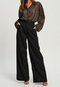 Pantaloni neri a gamba larga con vita alta, abbinati a una blusa trasparente a maniche lunghe con motivo, dotata di bottoni e colletto.