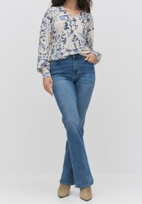Helle, gemusterte Bluse mit langen Ärmeln und V-Ausschnitt, kombiniert mit blauen Schlagjeans und beigen Stiefeletten. Glattes Material und entspannte Passform.