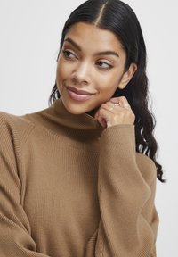 Rjava rebrasta turtleneck pulover z ohlapnim krojem. Ima zapleteno teksturo in dolge rokave. Model se z eno roko dotika ovratnika.