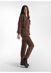 Bruine sweat-outfit met een losse sweatshirt met ronde hals en verlaagde schouders, gecombineerd met bijpassende taps toelopende broek. Gecombineerd met zwarte sneakers.