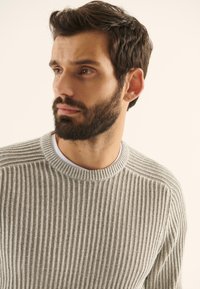 Jersey de color gris claro con relieve y un cuello redondo, usado sobre una camisa blanca, con sutiles detalles de costura.
