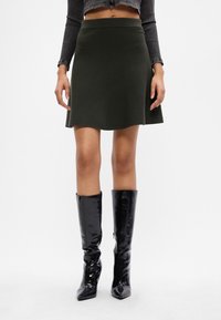 Žena oblečená v tmavozelenej rozšírenej sukni, čiernych lesklých čižmách po kolená a skrátenom pruhovanom svete na bielom pozadí.