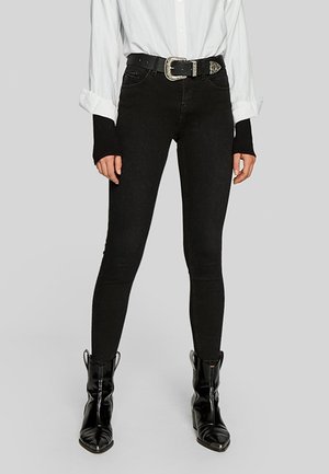 Femme portant un jean skinny noir, une ceinture style western noire, et des bottines noires à bout pointu avec une chemise blanche à manches longues et des poignets noirs.