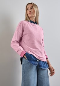 Pullover in maglia rosa con maniche lunghe e polsini a coste, indossato sopra una camicia in denim blu. Il pullover ha una vestibilità morbida e un tessuto texturizzato.