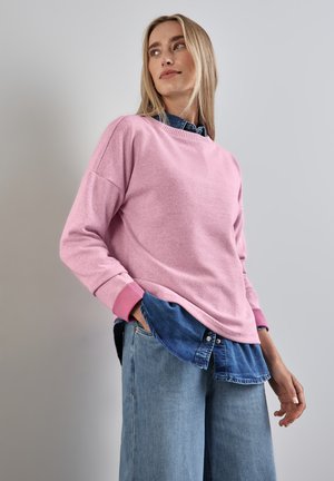 Pullover in maglia rosa con maniche lunghe e polsini a coste, indossato sopra una camicia in denim blu. Il pullover ha una vestibilità morbida e un tessuto texturizzato.
