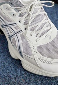 Close-up de um sapatilho desportivo cinza claro com um intrincado design de malha e atacadores, sobre um fundo de tapete azul texturizado.