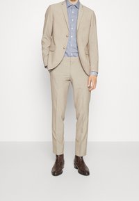 Beige linneskjorta med två knappar, skräddarsydd passform. Under den en blå och vit rutmönstrad skjorta. Bruna mönstrade läderstövlar.