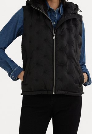 Femme portant un gilet matelassé noir avec fermeture éclair à l'avant sur une chemise en denim bleu, mains dans les poches du gilet, associé à un pantalon noir.